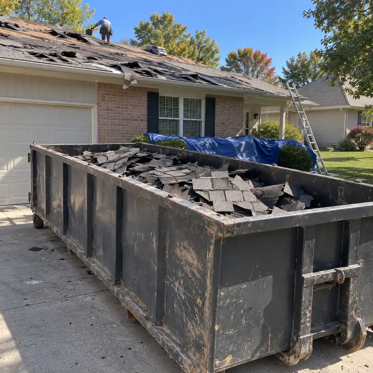 Old asphalt shingles from a roof tear-off piled in a dumpster beside a house